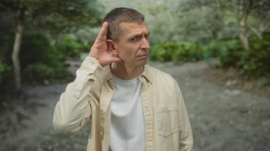 Serin bir açık hava parkında dikkatlice dinleyen bir adam. Etrafı yemyeşil, elleri kulaklarına kenetlenmiş, sakin bir günde günlük giysiler giymiş..