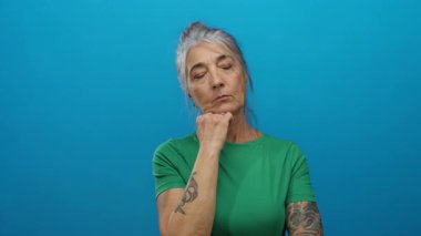 Gri saçlı ve dövmeli bir kadın yeşil gömlekli, parlak mavi bir arkaplan üzerinde düşünceli bir şekilde kafa yoruyor, özgüveni ve özgüvenini somutlaştırıyor..