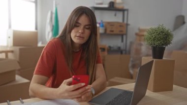 Oturma odasındaki kadın telefon ve dizüstü bilgisayarında taşınan kutuların arasında karton kutularla yeni bir ev ortamı ve bitki renklendirme ve teknoloji gösteriyor..