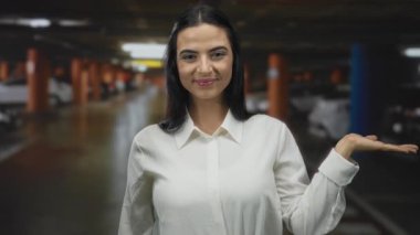Kapalı otoparkta elini açık bir şekilde sunan, arka planda beyaz gömlekli, özgüvenini ve dikkatini ifade eden arabaları olan bir kadın..