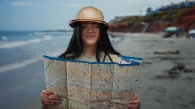 Deniz kenarını haritayla keşfeden bir kadın, sırt çantası ve şapka takan, parlak bir gökyüzünün altında manzaralı deniz manzaralı bir sahilde tatilin tadını çıkaran bir kadın..