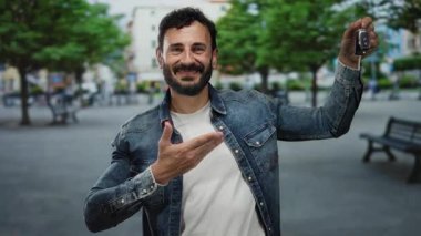 Sokakta elinde araba anahtarı olan İspanyol bir adam yeşil ağaçların altında kot ceket ve beyaz gömlek giyen neşeli bir gülümsemeyle işaret ediyor..
