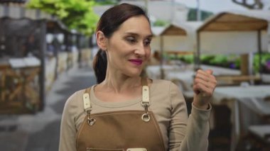 Restoran terasında gülümseyen önlüklü bir kadın canlı sokak dekorasyonu ve hoş bir atmosfer sergileyerek şehir ortamında misafirperverliği ön plana çıkarıyor..