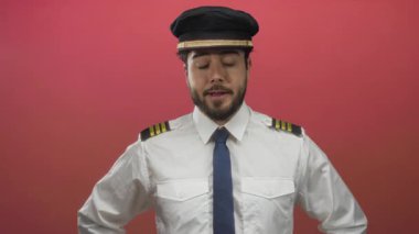 Genç İspanyol sakallı üniformalı adam canlı kırmızı bir arka plana karşı güvenle poz veriyor, bir pilot kılığındaki profesyonelliği ve otoriteyi temsil ediyor..