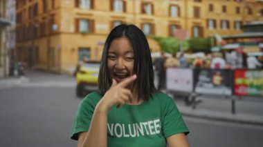 Yeşil gönüllü tişörtlü genç Çinli kadın hareketli bir şehir caddesinde enerjik bir şekilde gülümsüyor ve el kol hareketi yapıyor..