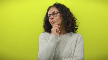 Süveter giyen kıvırcık saçlı İspanyol kadın parlak sarı bir arkaplan karşısında düşünceli bir tavırla düşünceli bir şekilde yansıtıcı ve izole bir sahne yaratıyor..