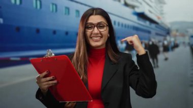 Limanda pano ile gülümseyen, gözlüklü, arka planda insanlarla birlikte büyük teknenin yanında duran, canlı bir ortamda kendine güvenen ve profesyonellik sergileyen bir kadın..