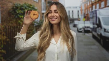 Genç sarışın kadın işlek şehir caddesinde göz hizasında jöleli donut tutuyor; keyif düşkünü hoşgörü zevki.