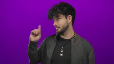 Sakallı, genç İspanyol bir adam telefon ediyor ve parlak bir mor arka plana işaret ediyor. Günlük ceket ve koyu gömlek giyiyor..