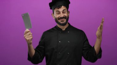 Siyah üniformalı İspanyol şef elinde satırla canlı mor arka planda aşçılık hevesine gülümsüyor..