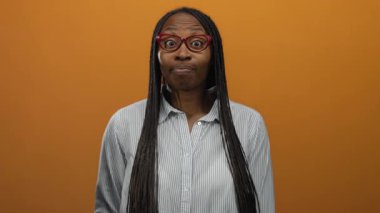 Gözlük takan bir kadın turuncu bir duvara karşı sessiz hareketler yapıyor, izole bir ortamda çizgili bir gömlekle gizemli bir ruh halini ve ifadeyi gösteriyor..