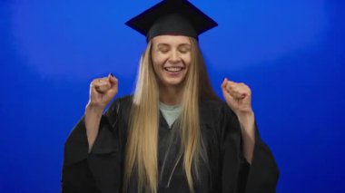 Mezuniyeti kep ve cüppeyle kutlayan kadın, mavi arkaplanın önünde neşeli bir ifadeyle duruyor ve el kaldırıyor, izole edilmiş bir görüntü..