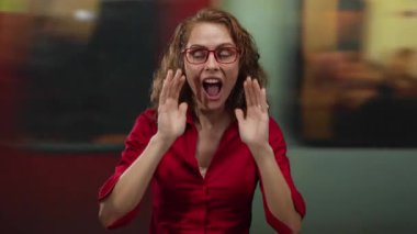 Kırmızı gömlekli ve gözlüklü bir kadın canlı şehir ortamında elleri ağızında bağırıyor.