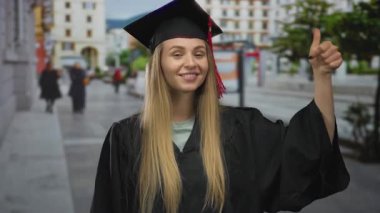 Mezuniyet üniformalı bir kadın güneşli bir günde şehir sokağında dikilip arka planda binalarla baş parmak gösteriyor..