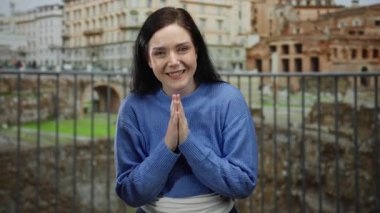 Esmer saçlı bir kadın güzel tarihi Roma harabelerinin önünde huzur içinde meditasyon yapıyor mavi bir kazak giyiyor ve elleri dua pozisyonunda gülümsüyor..