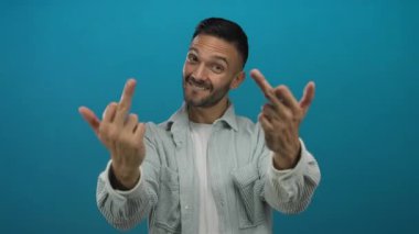 Sakallı genç adam, neşeli ve alışılmadık bir ifadeyi yakalayarak, parlak mavi bir arka plana karşı müstehcen bir jest yaparken gülümsüyor..