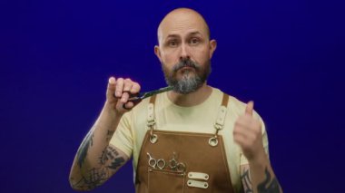 Sakallı bir erkek berber. Mavi arka planda makas tutuyor. Dövmeli, sarı tişörtlü ve deri önlüklü..