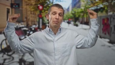 Bir adam şehir caddesinde anlamlı bir şekilde el kol hareketi yapıyor, günlük gömlekler giyiyor, açık havada ağaçların ve bisikletlerin olduğu bir yerde iletişim kuruyor ve ifade ediyor..