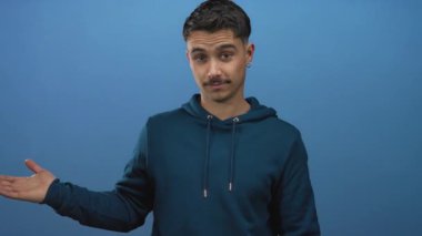 Kapüşonlu genç İspanyol bir adam başparmağıyla izole edilmiş mavi bir arka plana doğru hareketler yapıyor, kendinden emin bir ifadeyle onay ve yön veriyor..