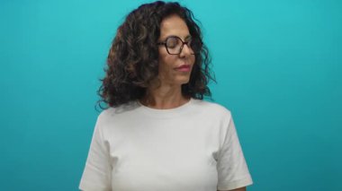 Gözlüklü İspanyol kadın yeşil bir duvara dayanıyor, beyaz gömlek giyerken şaşırdığını ifade ediyor, soyutlanmış ve orta yaşlı..