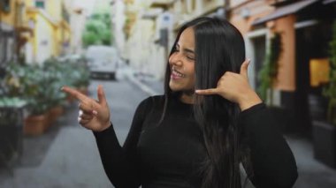 Açık havada el kol hareketi yapan genç bir kadın canlı şehir sokak ortamında neşeli bir ifadeyle beni arıyor.