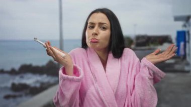 Pembe bornoz giymiş bir kadın deniz kenarında gezinirken elinde deniz arka planında şaşkın bir ifadeyle elektrikli bir diş fırçası tutuyor..