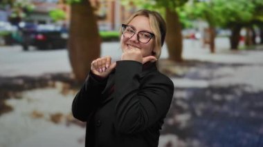 Güneşli bir günde, gülümseyen ve işaret eden bir kadın gözlük ve siyah bir ceket takıyor..