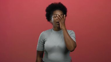 Yüzü koyun gülen bir kadın, izole edilmiş kırmızı arka plana karşı duruyor, neşe ve şaşkınlık ifade ediyor, afro saç stili, gri gömlek, genç Afro-Amerikan.