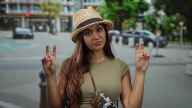 Hasır şapkalı kadın iki eliyle şehir sokağında barış işareti yapıyor. Kameralar sallanıyor, gençlik ruhu sergiliyor..