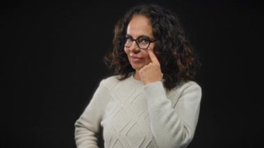 Kıvırcık saçlı bir kadın, siyah arka planda dikilirken, beyaz bir kazak giyerken, odaklanmış ve düşünceli bir ifade yaratırken, gözünü işaret ediyor..