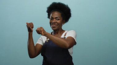 Önlükle gülümseyen kadın başparmakları geriye doğru izole edilmiş mavi arka plana bakıyor