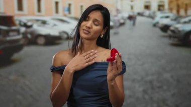 Kadın şehir caddesinde nişan yüzüğü tutuyor, duygularını ifade ediyor, güneşli şehir ortamında bulanık araba ve binalara karşı evlenme teklifi yapıyor..