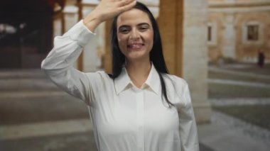 Eski bir üniversite avlusunda gülümserken gözlerini kapayan genç bir kadın mimari arka planda neşeli bir an yaşatıyor..