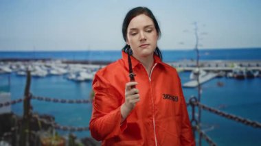 Turuncu hapishane tulumlu kadın arka planda teknelerle limanda bıçak tutuyor, gergin bir ifade sergiliyor..