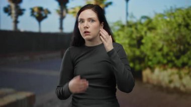 Sokakta gezen kadın şaşırmış görünüyor. Elleri havada, güneşli bir arka planda, güneşli bir günde ağaçlar ve şehir manzarası eşliğinde günlük siyah kıyafetler giyiyor..