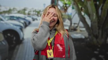 Şehirde bir kadın sokakta gönüllü olarak gülerken bulutlu gökyüzünün altında kırmızı yelek ve sarı rozet takıyor..