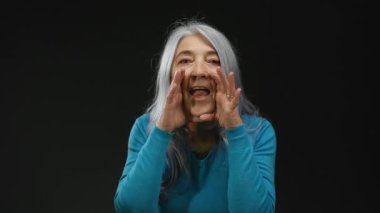 Gri saçlı kadın, siyah arka plana karşı anlamlı bir şekilde konuşuyor, mavi kazak giyiyor, izole bir iletişim ve ifade sahnesinde enerji ve duygu taşıyor..