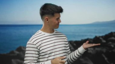 Çizgili tişörtlü genç İspanyol bir adam kumsalda durmuş avucunu işaret ediyor ve sakin bir deniz manzarasına başparmak işareti yapıyor.