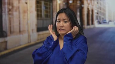 Şehir sokaklarında mavi tişörtlü bir kadın, elleriyle endişeli görünüyor, şehrin arka planında kentsel bir kaygı olduğunu gösteriyor..