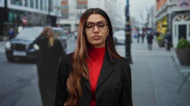 Kırmızı bluzlu ve siyah ceketli kadın, arka planda bulanık yayalar ve araçlarla yoğun şehir caddesinde güven içinde duruyor.