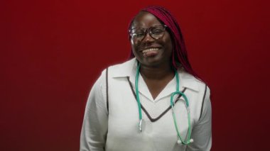 Kırmızı duvara karşı steteskop takan, parmaklarıyla buruna dokunarak gülümseyen Afrikalı Amerikalı kadın, profesyonel sağlık mutluluğunu aktarıyor..