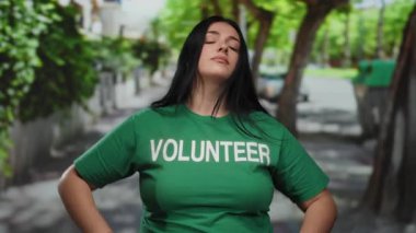 Yeşil gönüllü gömlekli kadın, güneşli şehir caddesinde kameraya bakıyor. Kendinden emin ve gururlu. Şehir gönüllülüğünü ve toplum ruhunu gösteriyor..