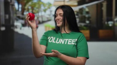 Şehir dışında yeşil gönüllü tişörtü giyen, elinde kırmızı bir kalp tutan, gülen bir kadın. Bu, hayır işi ve gündüz vakti toplumsal katılımı simgeliyor..