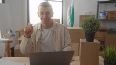 Oturma odasındaki adam video görüşmesi sırasında kutularla çevrili, elinde anahtarlar, masada dizüstü bilgisayarla, yeni ev ortamını ve heyecanı simgeliyor..