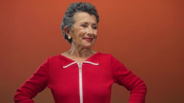 Gri saçlı, turuncu arka planda canlı kırmızı elbisesiyle güvenle gülümseyen son sınıf öğrencisi bir kadın. Bir stüdyo ortamında zarafet ve duruş sergiliyor..