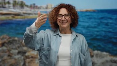 İspanyol orta yaşlı, kıvırcık esmer saçlı, güneşli bir sahilde gülümseyen, gözlüklü ve rahat gömlekli bir kadın..