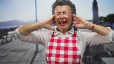 Deniz kenarında kulaklarını örten önlüklü, gri saçlı yaşlı bir kadın. Gün boyunca plaj ve okyanus manzarasıyla çevrili endişeli bir ifadeyle..