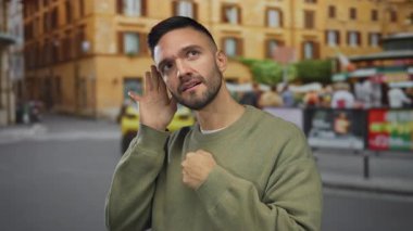 Kentsel sokaktaki Latin kökenli genç adam el pençe divan dururken şehir merkezindeki arka planda bulanık binalar ve pankartlarla süveter giyiyor..