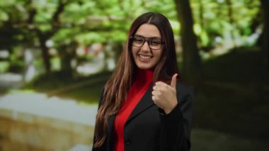 Gözlüklü bir kadın, yemyeşil bir parkta başparmağını kaldırarak gülümsüyor. Gündüz vakti açık havada mutluluk ve güveni gösteriyor..