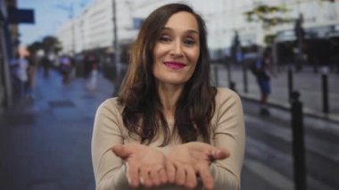 İspanyol kadın kalabalık bir sokak sahnesinde avuçlarını kameraya doğru açıyor; davetiye açıklığı sıcaklığı nezaketi.